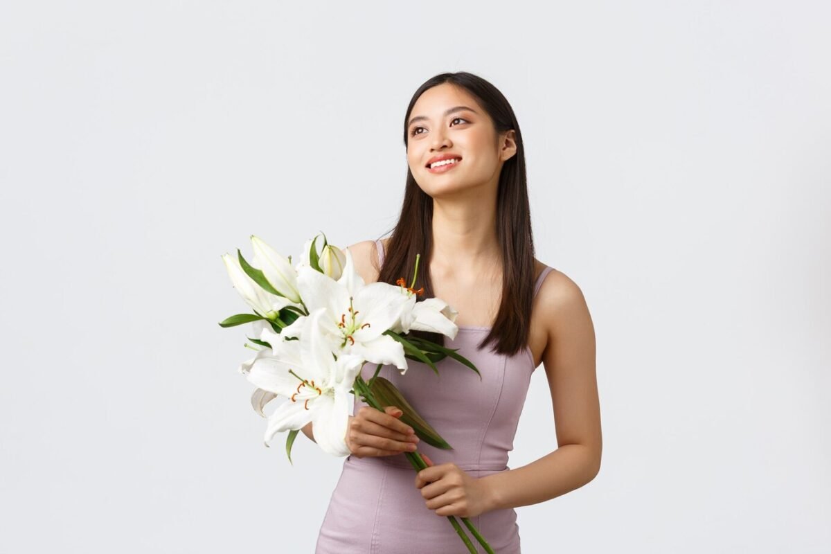 happy-smiling-asian-woman-stylish-dress-looking-upper-left-corner-holding-bouquet-lilies_1258-28262